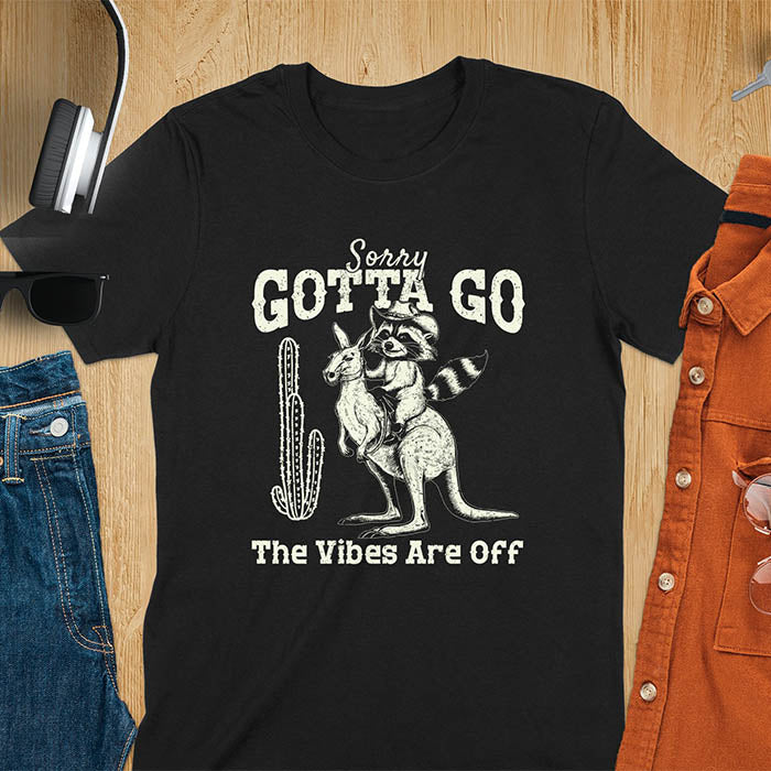 A t-shirt with a raccoon wearing a cowboy hat riding a kangaroo. Text: Sorry GOTTA GO above and The Vibes Are Off below. This humorous tee-shirt features a funny gen z meme and sarcastic meme shirt, a weird graphics shirt. A funny retro graphic and vintage graphic tee with rodeo cowboy elements. A silly shirt or funny unhinged shirt, an animal person gift or kangaroo gifts. A raccoon t shirt or mom gift alternative.