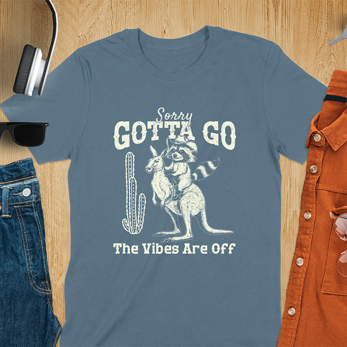 A t-shirt with a raccoon wearing a cowboy hat riding a kangaroo. Text: Sorry GOTTA GO above and The Vibes Are Off below. This humorous tee-shirt features a funny gen z meme and sarcastic meme shirt, a weird graphics shirt. A funny retro graphic and vintage graphic tee with rodeo cowboy elements. A silly shirt or funny unhinged shirt, an animal person gift or kangaroo gifts. A raccoon t shirt or mom gift alternative.