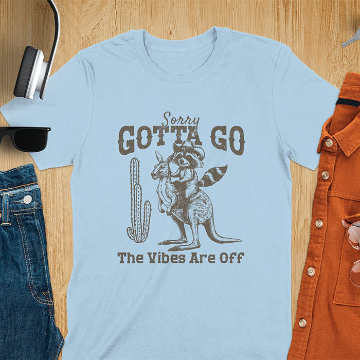 A t-shirt with a raccoon wearing a cowboy hat riding a kangaroo. Text: Sorry GOTTA GO above and The Vibes Are Off below. This humorous tee-shirt features a funny gen z meme and sarcastic meme shirt, a weird graphics shirt.  A funny retro graphic and vintage graphic tee with rodeo cowboy elements. A silly shirt or funny unhinged shirt, an animal person gift or kangaroo gifts. A raccoon t shirt or mom gift alternative.