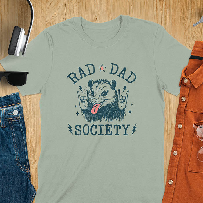 A t-shirt with an opossum sticking its tongue out and giving rock-on hand signs. Text: "RAD DAD" above and "SOCIETY" below. This rad dad society tee is for the cool dad club. A dad humor gift and cool dad tee-shirt, a daddie's t shirt or dad birthday shirt. A first father's day or fathers day gifts, a new daddy gift or gift for daddie. A pet dad gift for a husband outfit.