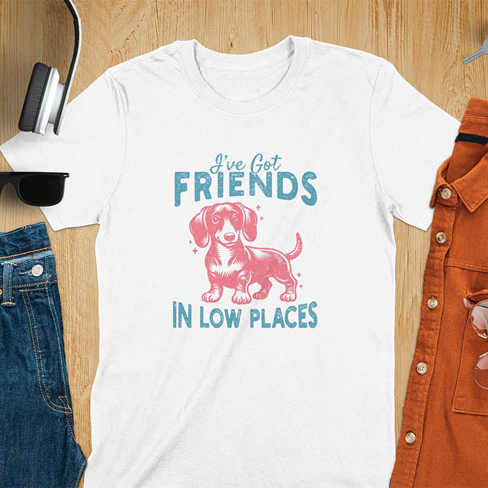 This T-shirt features a vintage-style line art of a happy dachshund looking straight at you with its tail held high. The top text reads, I’ve got friends, and below it says, in low places. Overall in a retro color palette with a distressed texture.