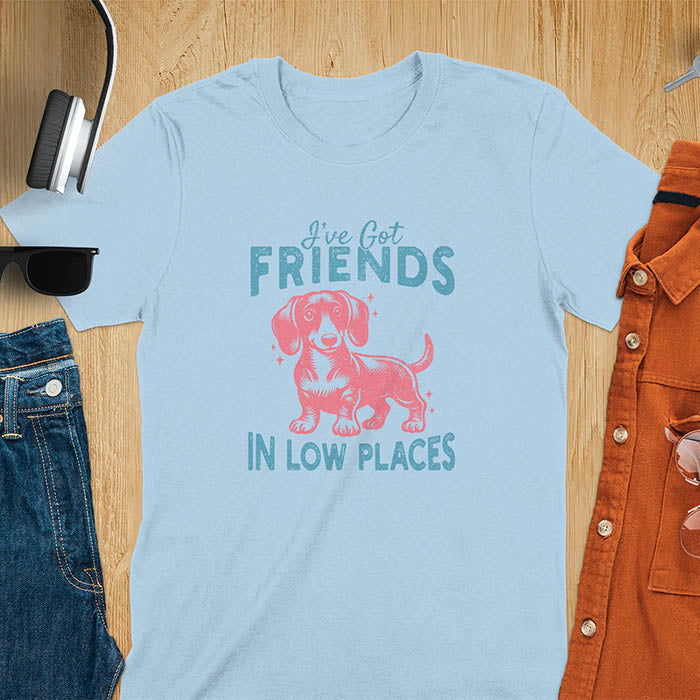 This T-shirt features a vintage-style line art of a happy dachshund looking straight at you with its tail held high. The top text reads, I’ve got friends, and below it says, in low places. Overall in a retro color palette with a distressed texture.