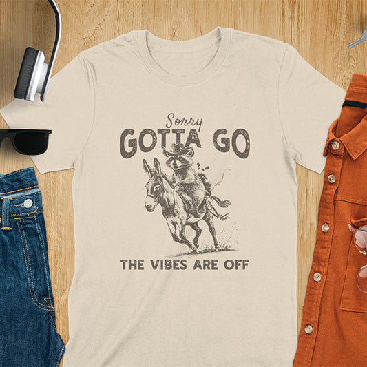A t-shirt with a raccoon wearing a cowboy hat riding a donkey, with an opossum seated behind. Text: Sorry GOTTA GO above and THE VIBES ARE OFF below. This humorous T-shirt features a funny donkey and opossum possum, a weird graphics shirt. A sarcastic meme shirt and funny Gen Z meme, with rodeo cowboy elements. This vintage graphic is one of those shirts that go hard, an animal person gift or raccoon person gift. A funny unhinged shirt or mom gift alternative.