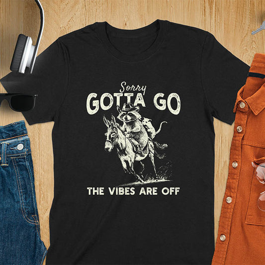 A t-shirt with a raccoon wearing a cowboy hat riding a donkey, with an opossum seated behind. Text: Sorry GOTTA GO above and THE VIBES ARE OFF below. This humorous T-shirt features a funny donkey and opossum possum, a weird graphics shirt. A sarcastic meme shirt and funny Gen Z meme, with rodeo cowboy elements. This vintage graphic is one of those shirts that go hard, an animal person gift or raccoon person gift. A funny unhinged shirt or mom gift alternative.