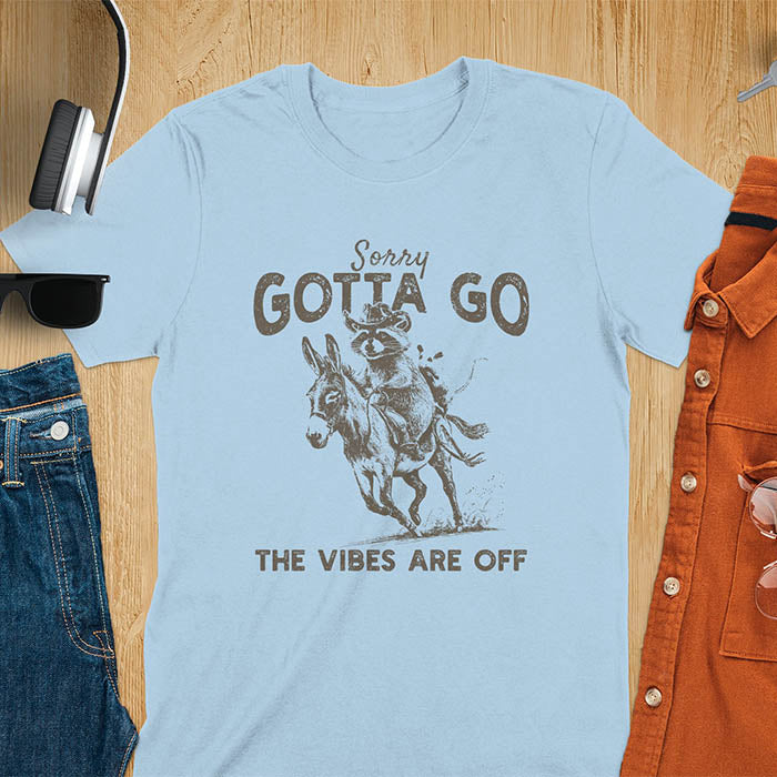 A t-shirt with a raccoon wearing a cowboy hat riding a donkey, with an opossum seated behind. Text: Sorry GOTTA GO above and THE VIBES ARE OFF below. This humorous T-shirt features a funny donkey and opossum possum, a weird graphics shirt. A sarcastic meme shirt and funny Gen Z meme, with rodeo cowboy elements. This vintage graphic is one of those shirts that go hard, an animal person gift or raccoon person gift. A funny unhinged shirt or mom gift alternative.