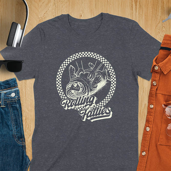A T-shirt design featuring a detailed illustration of a chubby basset hound being rolled by two hands, surrounded by a checkered circle. The text Rolling Fatties is written in a stylized font below the image. Retro style with distressed texture