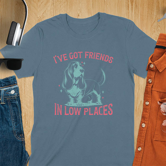 This retro dog shirt features a vintage dog tee design of a basset hound standing proudly, tail high, and gazing to the side. Text reads “I’ve got friends” above and “in low places” below. A distressed texture adds charm to the shirt.