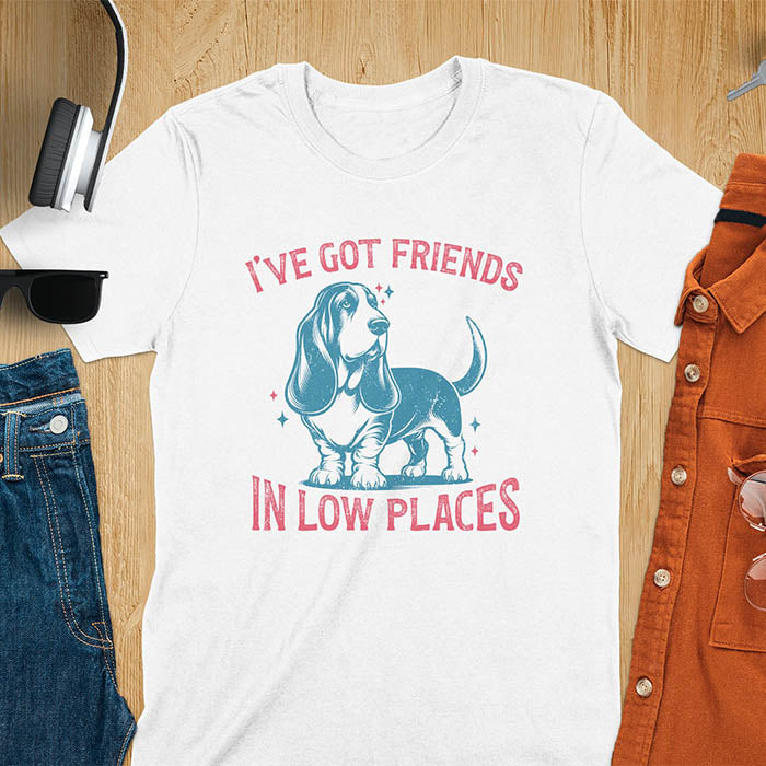 This retro dog shirt features a vintage dog tee design of a basset hound standing proudly, tail high, and gazing to the side. Text reads “I’ve got friends” above and “in low places” below. A distressed texture adds charm to the shirt.