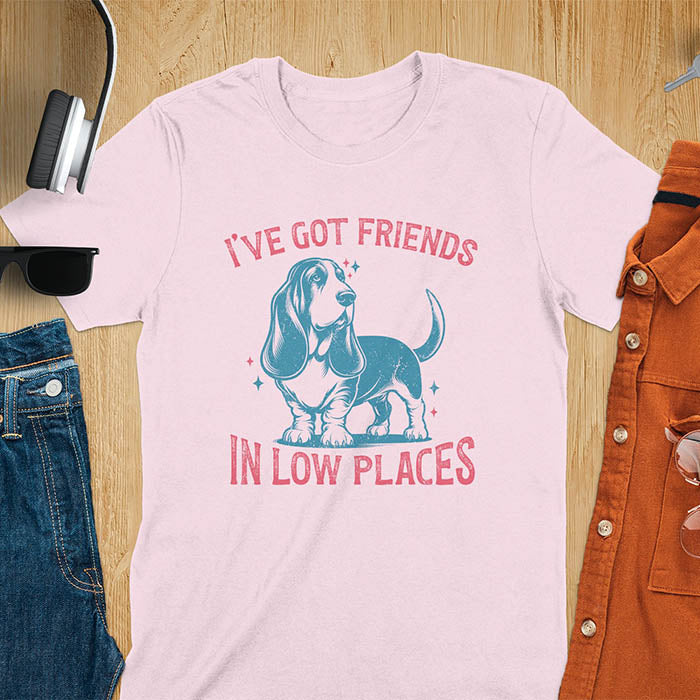 This retro dog shirt features a vintage dog tee design of a basset hound standing proudly, tail high, and gazing to the side. Text reads “I’ve got friends” above and “in low places” below. A distressed texture adds charm to the shirt.