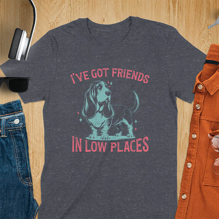 This retro dog shirt features a vintage dog tee design of a basset hound standing proudly, tail high, and gazing to the side. Text reads “I’ve got friends” above and “in low places” below. A distressed texture adds charm to the shirt.