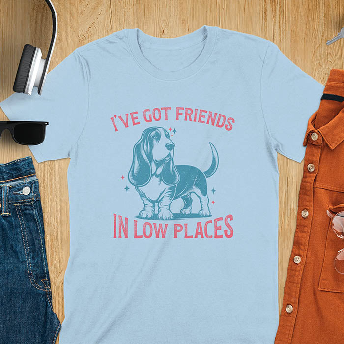 This retro dog shirt features a vintage dog tee design of a basset hound standing proudly, tail high, and gazing to the side. Text reads “I’ve got friends” above and “in low places” below. A distressed texture adds charm to the shirt.