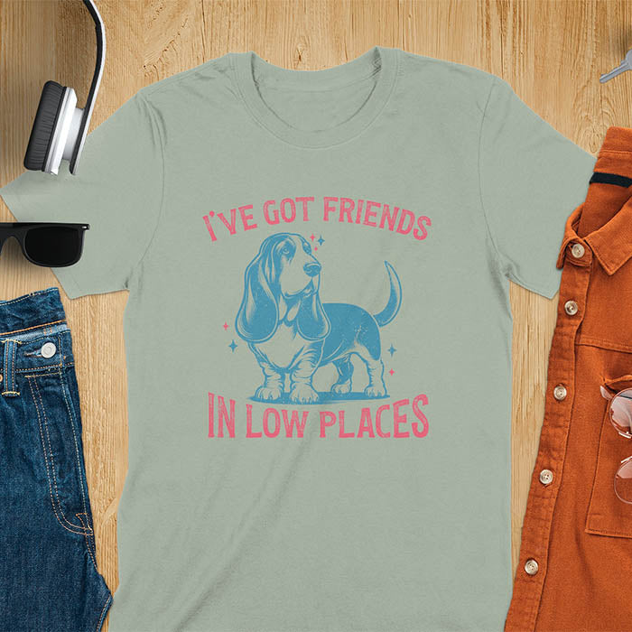 This retro dog shirt features a vintage dog tee design of a basset hound standing proudly, tail high, and gazing to the side. Text reads “I’ve got friends” above and “in low places” below. A distressed texture adds charm to the shirt.