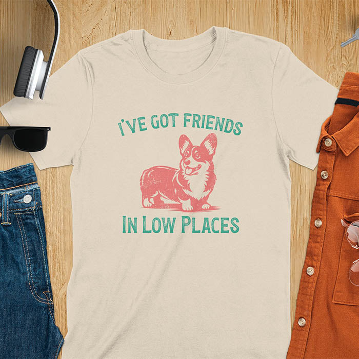 Our vintage-inspired design features a cute corgi silhouette with facial details. Text above it says 'I've Got Friends' and below it says ''In Low Places.' With its retro vibe and distressed texture, this tee adds a touch of nostalgia to your wardrobe.