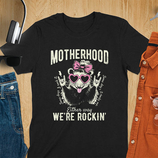 A t-shirt with an opossum wearing a bow and heart-shaped sunglasses, throwing rock-on hand gestures. Text: MOTHERHOOD above, Some days I rock it left, Some days it rocks me right, and Either way WE'RE ROCKIN below. Lightning bolt accents on the hands. This vintage mama tee or rocker mom shirt is a trending mama design with a mom saying. A snarky humor designs and unhinged shirts style, a mom gift hers or baby shower gift. A first time mom gift or part of mommy outfits, for rocking motherhood.