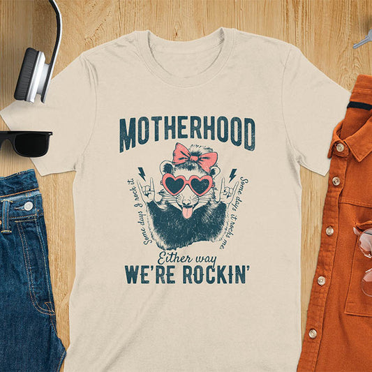 A t-shirt with an opossum wearing a bow and heart-shaped sunglasses, throwing rock-on hand gestures. Text: MOTHERHOOD above, Some days I rock it left, Some days it rocks me right, and Either way WE'RE ROCKIN below. Lightning bolt accents on the hands. This vintage mama tee or rocker mom shirt is a trending mama design with a mom saying. A snarky humor designs and unhinged shirts style, a mom gift hers or baby shower gift. A first time mom gift or part of mommy outfits, for rocking motherhood.