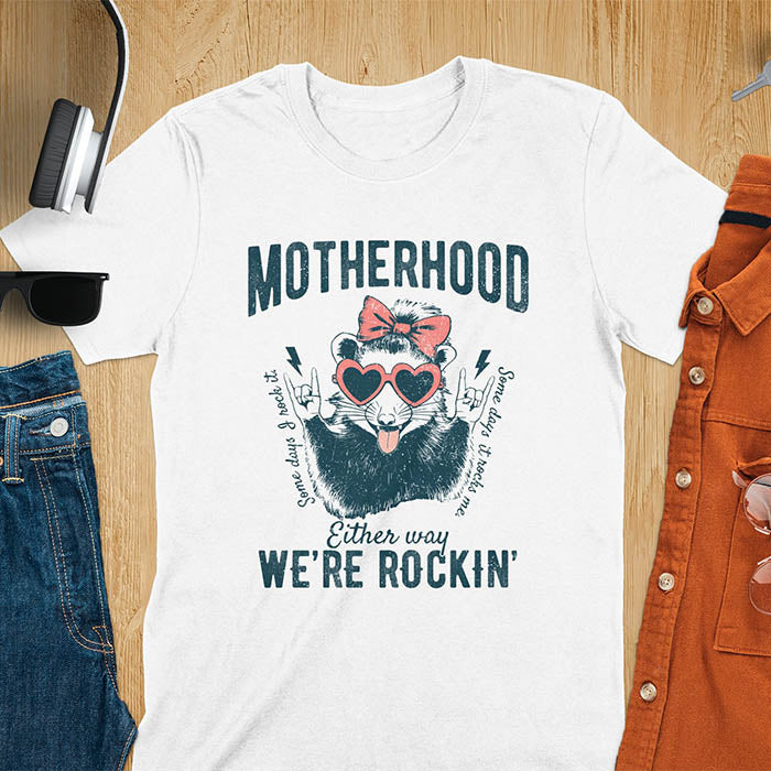 A t-shirt with an opossum wearing a bow and heart-shaped sunglasses, throwing rock-on hand gestures. Text: MOTHERHOOD above, Some days I rock it left, Some days it rocks me right, and Either way WE'RE ROCKIN below. Lightning bolt accents on the hands. This vintage mama tee or rocker mom shirt is a trending mama design with a mom saying. A snarky humor designs and unhinged shirts style, a mom gift hers or baby shower gift. A first time mom gift or part of mommy outfits, for rocking motherhood.