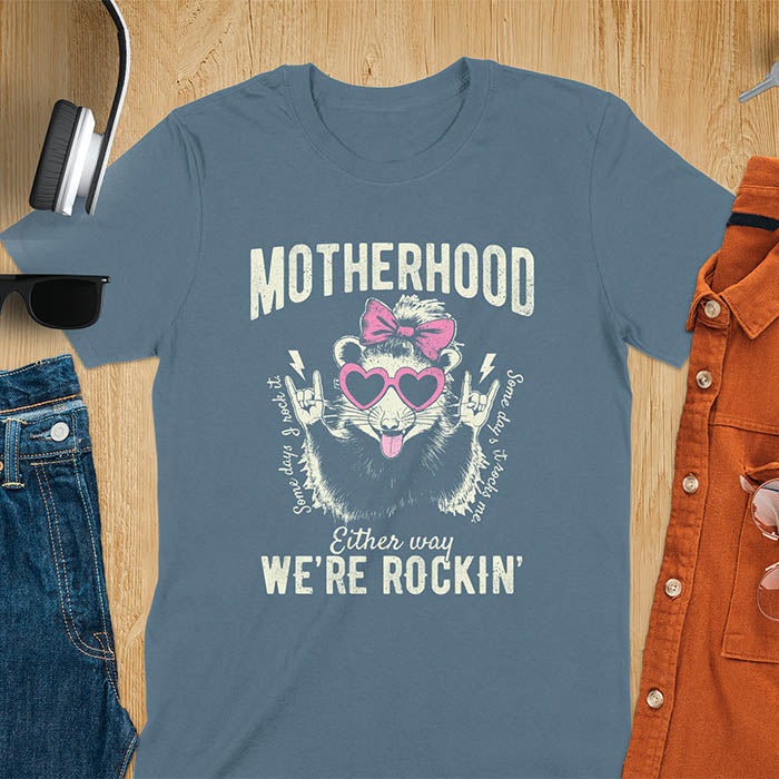 A t-shirt with an opossum wearing a bow and heart-shaped sunglasses, throwing rock-on hand gestures. Text: MOTHERHOOD above, Some days I rock it left, Some days it rocks me right, and Either way WE'RE ROCKIN below. Lightning bolt accents on the hands. This vintage mama tee or rocker mom shirt is a trending mama design with a mom saying. A snarky humor designs and unhinged shirts style, a mom gift hers or baby shower gift. A first time mom gift or part of mommy outfits, for rocking motherhood.