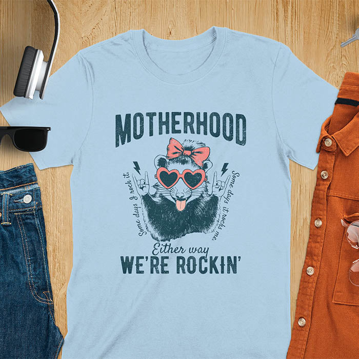 A t-shirt with an opossum wearing a bow and heart-shaped sunglasses, throwing rock-on hand gestures. Text: MOTHERHOOD above, Some days I rock it left, Some days it rocks me right, and Either way WE'RE ROCKIN below. Lightning bolt accents on the hands. This vintage mama tee or rocker mom shirt is a trending mama design with a mom saying. A snarky humor designs and unhinged shirts style, a mom gift hers or baby shower gift. A first time mom gift or part of mommy outfits, for rocking motherhood.