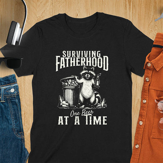 A t-shirt with a raccoon holding a beer next to a overflowing trash can. Text: "SURVIVING" above "FATHERHOOD" and "One Beer" above "AT A TIME" below. This Fatherhood shirt is a dad t shirt and dad humor gift. A Fathers day gift or Fathers day shirt, a dad birthday shirt and dad gift. A Funny father shirt and gift for daddie, a new daddy gift. Part of a husband outfit and Funny dad clothe, for surviving fatherhood.