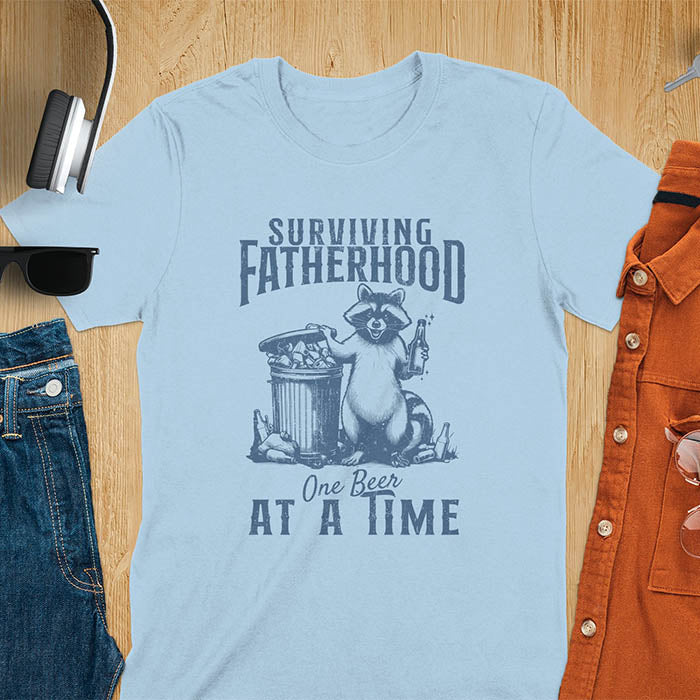 A t-shirt with a raccoon holding a beer next to a overflowing trash can. Text: "SURVIVING" above "FATHERHOOD" and "One Beer" above "AT A TIME" below. This Fatherhood shirt is a dad t shirt and dad humor gift. A Fathers day gift or Fathers day shirt, a dad birthday shirt and dad gift. A Funny father shirt and gift for daddie, a new daddy gift. Part of a husband outfit and Funny dad clothe, for surviving fatherhood.