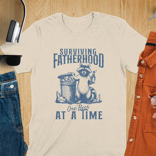 A t-shirt with a raccoon holding a beer next to a overflowing trash can. Text: "SURVIVING" above "FATHERHOOD" and "One Beer" above "AT A TIME" below. This Fatherhood shirt is a dad t shirt and dad humor gift. A Fathers day gift or Fathers day shirt, a dad birthday shirt and dad gift. A Funny father shirt and gift for daddie, a new daddy gift. Part of a husband outfit and Funny dad clothe, for surviving fatherhood.