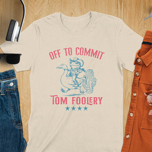 A t-shirt with a frog dressed as a rodeo cowboy riding a goose. Text: "Off To COMMIT" above and "TOM FOOLERY" below. This meme t shirt features a funny frog tee and silly funny goose, a weird graphics shirt. A funny genz meme and unhinged tee-shirt, with rodeo cowboy and cowgirl t shirt elements. A retro aesthetic and western style gift, an animal person gift. A mom gift alternative.
