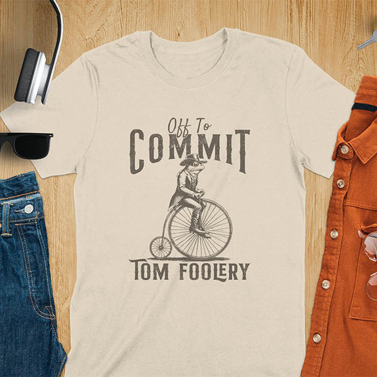 A t-shirt with a frog in vintage clothing riding a penny-farthing bicycle. Text: "Off To COMMIT" above and "TOM FOOLERY" below. This funny meme shirt is a weird graphics shirt with a retro aesthetic. A meme t shirt and unhinged t shirt, a gag gift idea. A funny frog tee and animal person gift, an animal lover gag. Touches on cottagecore weird and weirdcore memes. A mom gift alternative with a vintage graphic style.