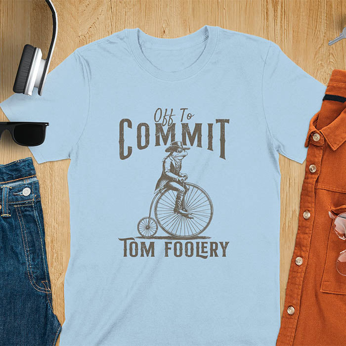 A t-shirt with a frog in vintage clothing riding a penny-farthing bicycle. Text: "Off To COMMIT" above and "TOM FOOLERY" below. This funny meme shirt is a weird graphics shirt with a retro aesthetic. A meme t shirt and unhinged t shirt, a gag gift idea. A funny frog tee and animal person gift, an animal lover gag. Touches on cottagecore weird and weirdcore memes. A mom gift alternative with a vintage graphic style.
