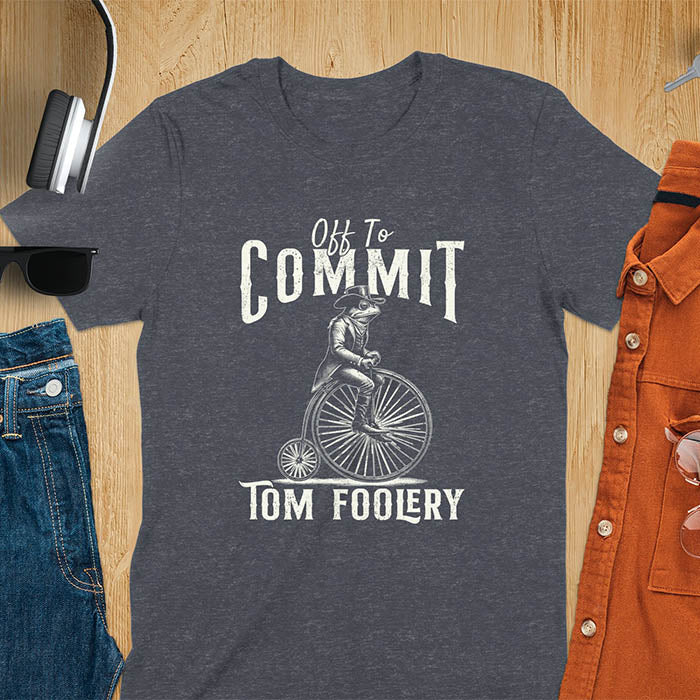 A t-shirt with a frog in vintage clothing riding a penny-farthing bicycle. Text: "Off To COMMIT" above and "TOM FOOLERY" below. This funny meme shirt is a weird graphics shirt with a retro aesthetic. A meme t shirt and unhinged t shirt, a gag gift idea. A funny frog tee and animal person gift, an animal lover gag. Touches on cottagecore weird and weirdcore memes. A mom gift alternative with a vintage graphic style.