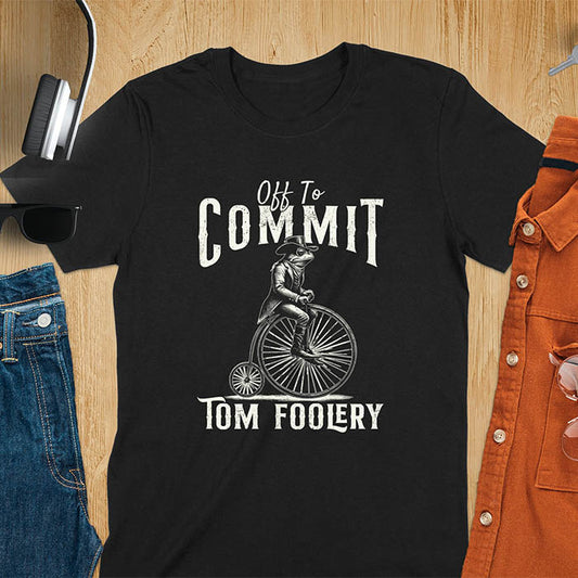 A t-shirt with a frog in vintage clothing riding a penny-farthing bicycle. Text: "Off To COMMIT" above and "TOM FOOLERY" below. This funny meme shirt is a weird graphics shirt with a retro aesthetic. A meme t shirt and unhinged t shirt, a gag gift idea. A funny frog tee and animal person gift, an animal lover gag. Touches on cottagecore weird and weirdcore memes. A mom gift alternative with a vintage graphic style.