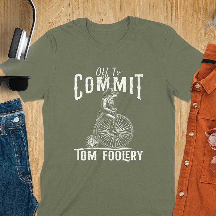 A t-shirt with a frog in vintage clothing riding a penny-farthing bicycle. Text: "Off To COMMIT" above and "TOM FOOLERY" below. This funny meme shirt is a weird graphics shirt with a retro aesthetic. A meme t shirt and unhinged t shirt, a gag gift idea. A funny frog tee and animal person gift, an animal lover gag. Touches on cottagecore weird and weirdcore memes. A mom gift alternative with a vintage graphic style.