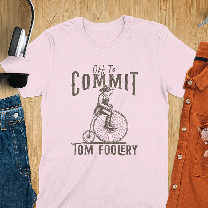 A t-shirt with a frog in vintage clothing riding a penny-farthing bicycle. Text: "Off To COMMIT" above and "TOM FOOLERY" below. This funny meme shirt is a weird graphics shirt with a retro aesthetic. A meme t shirt and unhinged t shirt, a gag gift idea. A funny frog tee and animal person gift, an animal lover gag. Touches on cottagecore weird and weirdcore memes. A mom gift alternative with a vintage graphic style.