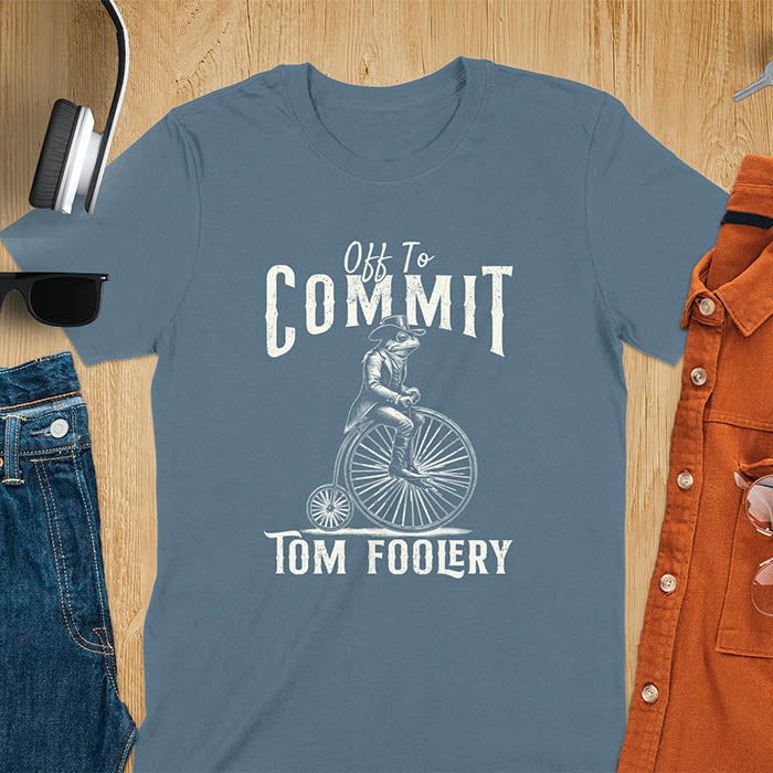 A t-shirt with a frog in vintage clothing riding a penny-farthing bicycle. Text: "Off To COMMIT" above and "TOM FOOLERY" below. This funny meme shirt is a weird graphics shirt with a retro aesthetic. A meme t shirt and unhinged t shirt, a gag gift idea. A funny frog tee and animal person gift, an animal lover gag. Touches on cottagecore weird and weirdcore memes. A mom gift alternative with a vintage graphic style.