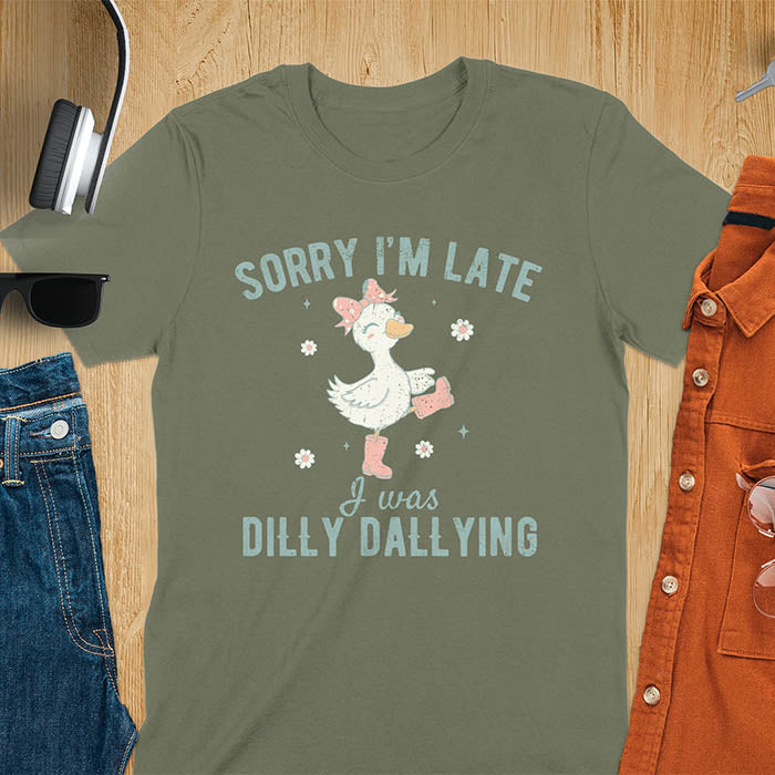 A t-shirt with a white goose wearing pink boots and a pink bow, surrounded by small flowers. Text: SORRY I'M LATE above and I was DILLY DALLYING below. This cute geese t shirt features goose cuteness and kawaiiness cuteness. A snarky humorous and sarcastic meme shirt about dilly dallying. A funny genz meme with funny animals and spring flowers. This silly funny goose design has vintage aesthetics and is an animal person gift or mom gift.