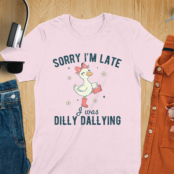 A t-shirt with a white goose wearing pink boots and a pink bow, surrounded by small flowers. Text: SORRY I'M LATE above and I was DILLY DALLYING below. This cute geese t shirt features goose cuteness and kawaiiness cuteness. A snarky humorous and sarcastic meme shirt about dilly dallying. A funny genz meme with funny animals and spring flowers. This silly funny goose design has vintage aesthetics and is an animal person gift or mom gift.
