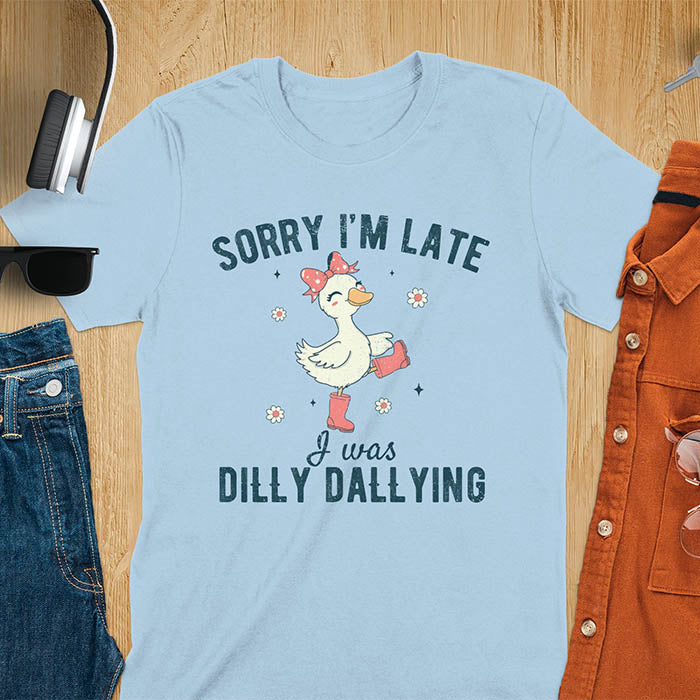 A t-shirt with a white goose wearing pink boots and a pink bow, surrounded by small flowers. Text: SORRY I'M LATE above and I was DILLY DALLYING below. This cute geese t shirt features goose cuteness and kawaiiness cuteness. A snarky humorous and sarcastic meme shirt about dilly dallying. A funny genz meme with funny animals and spring flowers. This silly funny goose design has vintage aesthetics and is an animal person gift or mom gift.