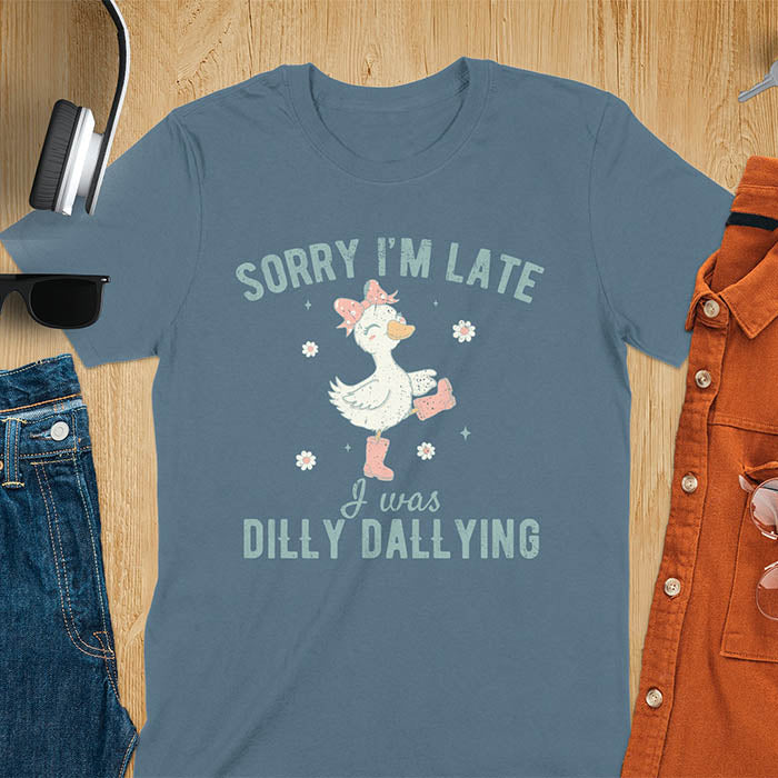 A t-shirt with a white goose wearing pink boots and a pink bow, surrounded by small flowers. Text: SORRY I'M LATE above and I was DILLY DALLYING below. This cute geese t shirt features goose cuteness and kawaiiness cuteness. A snarky humorous and sarcastic meme shirt about dilly dallying. A funny genz meme with funny animals and spring flowers. This silly funny goose design has vintage aesthetics and is an animal person gift or mom gift.