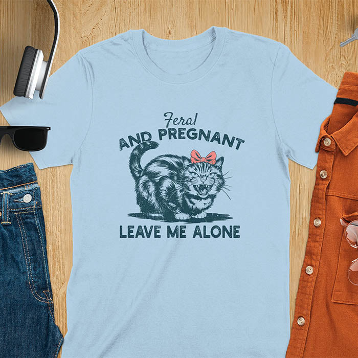 A t-shirt with a tabby cat with a grumpy expression and a pink bow. Text: Feral AND PREGNANT above and LEAVE ME ALONE below. This funny maternity tee is a feral cats shirt for a mom to be shirt. A pregnancy humor design for a pregnancy announce or sibling pregnancy. An expecting mom gift or mommy to be gift, a maternity apparel and pregnancy keepsake for an expecting baby during a feral pregnancy.