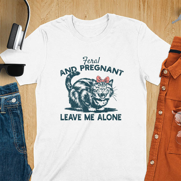 A t-shirt with a tabby cat with a grumpy expression and a pink bow. Text: Feral AND PREGNANT above and LEAVE ME ALONE below. This funny maternity tee is a feral cats shirt for a mom to be shirt. A pregnancy humor design for a pregnancy announce or sibling pregnancy. An expecting mom gift or mommy to be gift, a maternity apparel and pregnancy keepsake for an expecting baby during a feral pregnancy.