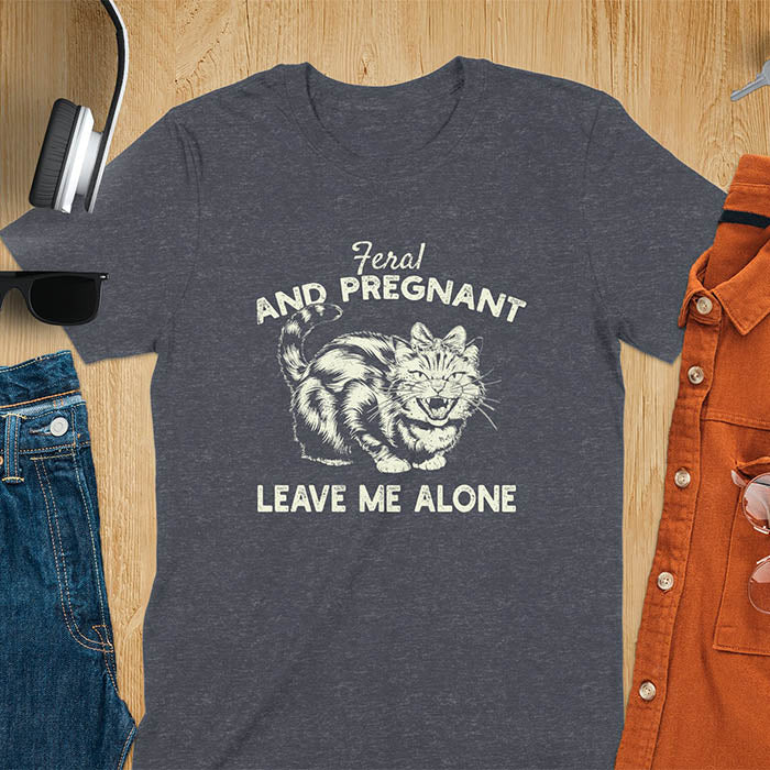 A t-shirt with a tabby cat with a grumpy expression and a pink bow. Text: Feral AND PREGNANT above and LEAVE ME ALONE below. This funny maternity tee is a feral cats shirt for a mom to be shirt. A pregnancy humor design for a pregnancy announce or sibling pregnancy. An expecting mom gift or mommy to be gift, a maternity apparel and pregnancy keepsake for an expecting baby during a feral pregnancy.