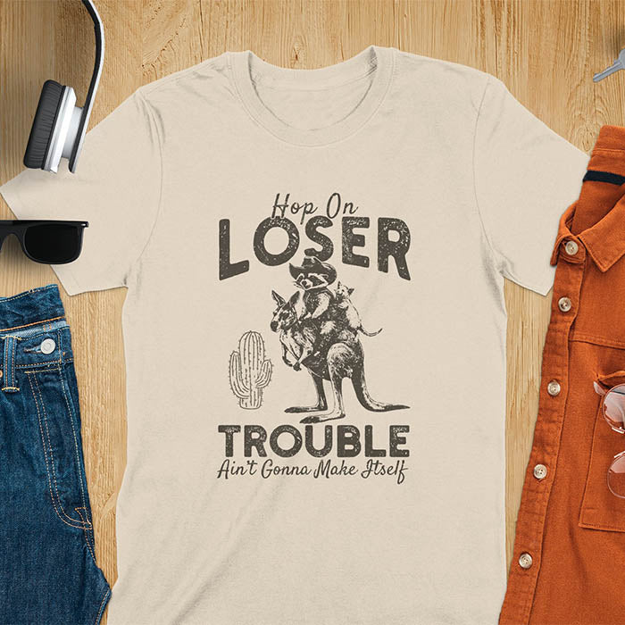 A t-shirt with a raccoon in cowboy attire riding a kangaroo, with an opossum behind. Text: "Hop On LOSER" above and "TROUBLE Ain't Gonna Make Itself" below. This meme t shirt features a funny raccoon (a cute trash panda) dressed as wild west cowboys riding with an opossum possum. A weird graphics shirt with a retro aesthetic, a sarcastic meme shirt and funny genz meme. An animal person gift for a cuteness lover, tapping into weirdcore memes. A mom Gift alternative.
