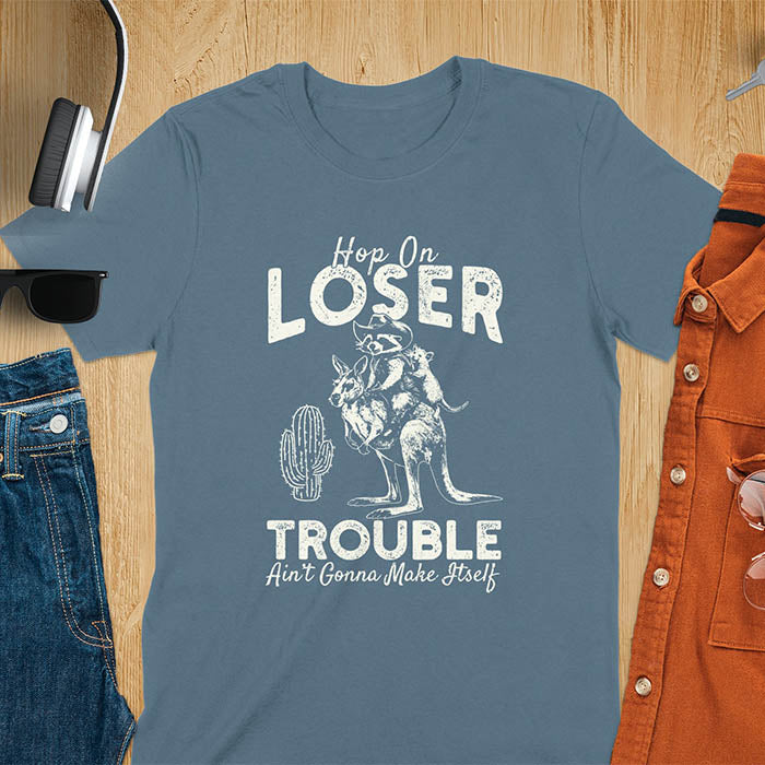 A t-shirt with a raccoon in cowboy attire riding a kangaroo, with an opossum behind. Text: "Hop On LOSER" above and "TROUBLE Ain't Gonna Make Itself" below. This meme t shirt features a funny raccoon (a cute trash panda) dressed as wild west cowboys riding with an opossum possum. A weird graphics shirt with a retro aesthetic, a sarcastic meme shirt and funny genz meme. An animal person gift for a cuteness lover, tapping into weirdcore memes. A mom Gift alternative.