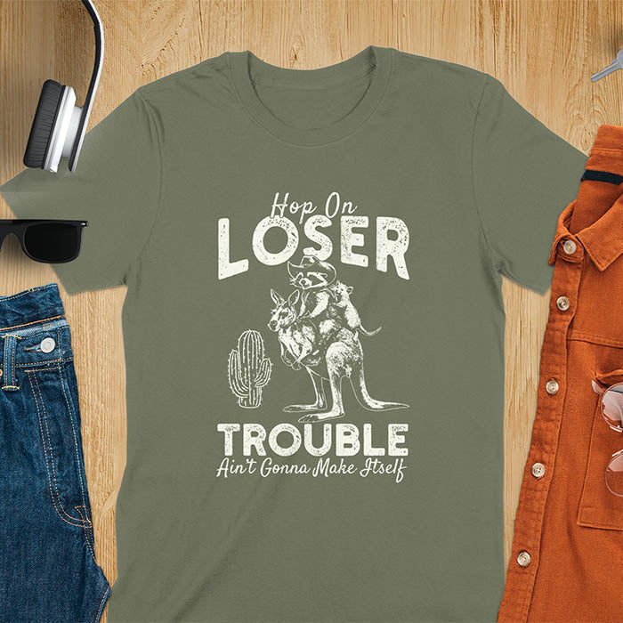 A t-shirt with a raccoon in cowboy attire riding a kangaroo, with an opossum behind. Text: "Hop On LOSER" above and "TROUBLE Ain't Gonna Make Itself" below. This meme t shirt features a funny raccoon (a cute trash panda) dressed as wild west cowboys riding with an opossum possum. A weird graphics shirt with a retro aesthetic, a sarcastic meme shirt and funny genz meme. An animal person gift for a cuteness lover, tapping into weirdcore memes. A mom Gift alternative.