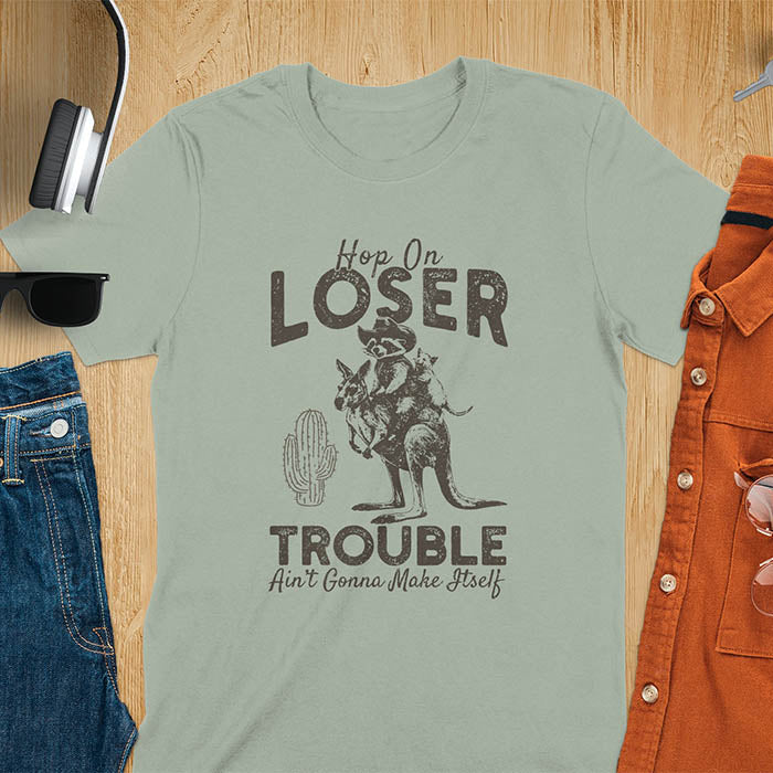 A t-shirt with a raccoon in cowboy attire riding a kangaroo, with an opossum behind. Text: "Hop On LOSER" above and "TROUBLE Ain't Gonna Make Itself" below. This meme t shirt features a funny raccoon (a cute trash panda) dressed as wild west cowboys riding with an opossum possum. A weird graphics shirt with a retro aesthetic, a sarcastic meme shirt and funny genz meme. An animal person gift for a cuteness lover, tapping into weirdcore memes. A mom Gift alternative.