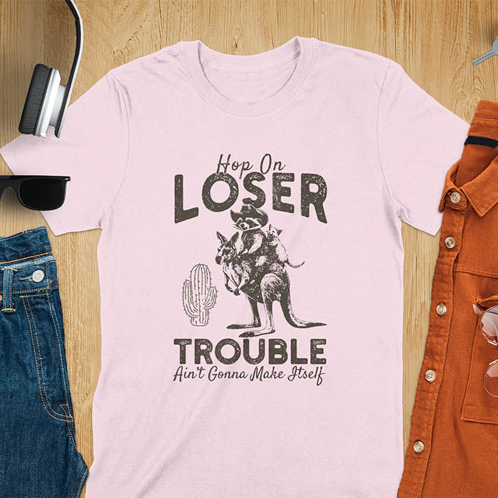 A t-shirt with a raccoon in cowboy attire riding a kangaroo, with an opossum behind. Text: "Hop On LOSER" above and "TROUBLE Ain't Gonna Make Itself" below. This meme t shirt features a funny raccoon (a cute trash panda) dressed as wild west cowboys riding with an opossum possum. A weird graphics shirt with a retro aesthetic, a sarcastic meme shirt and funny genz meme. An animal person gift for a cuteness lover, tapping into weirdcore memes. A mom Gift alternative.