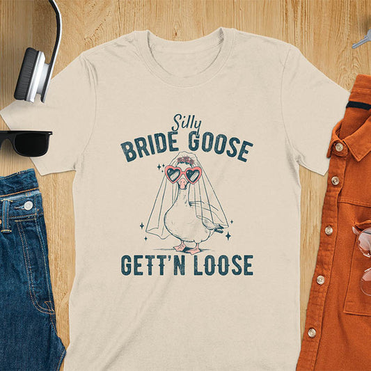 A t-shirt with a goose dressed as a bride with heart sunglasses and a veil. Text: "Silly BRIDE GOOSE" above and "GETT'N LOOSE" below. This funny bride shirt features a silly goose for getting ready bridal. A bride crewneck and engaged gift, a fiancé gift for honeymoon outfit or honeymoon gifts. A married couple shirt or new bride shirt, a fun engagement announce idea. A couple's tee-shirt and funny goose gift.