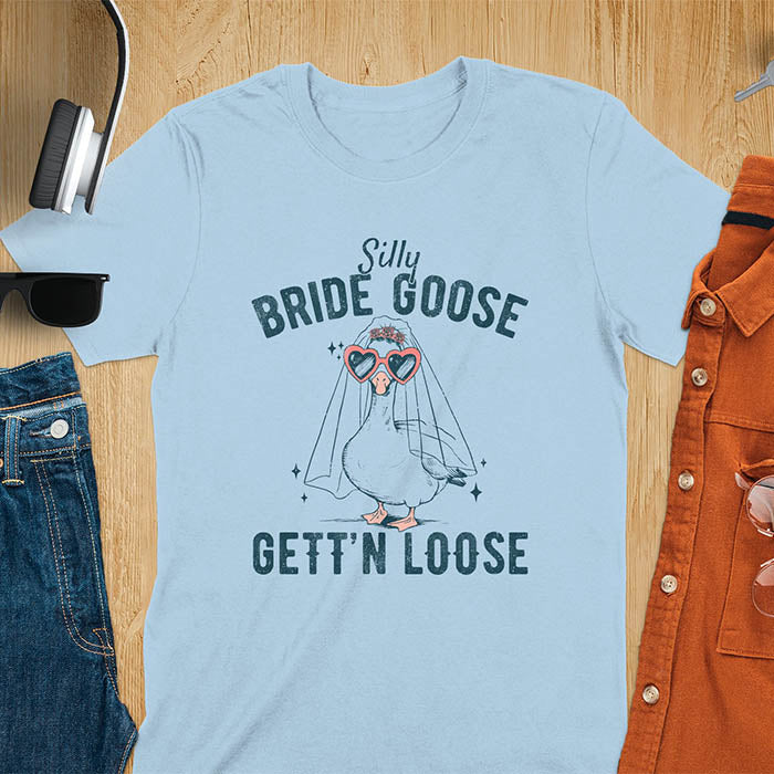 A t-shirt with a goose dressed as a bride with heart sunglasses and a veil. Text: "Silly BRIDE GOOSE" above and "GETT'N LOOSE" below. This funny bride shirt features a silly goose for getting ready bridal. A bride crewneck and engaged gift, a fiancé gift for honeymoon outfit or honeymoon gifts. A married couple shirt or new bride shirt, a fun engagement announce idea. A couple's tee-shirt and funny goose gift.