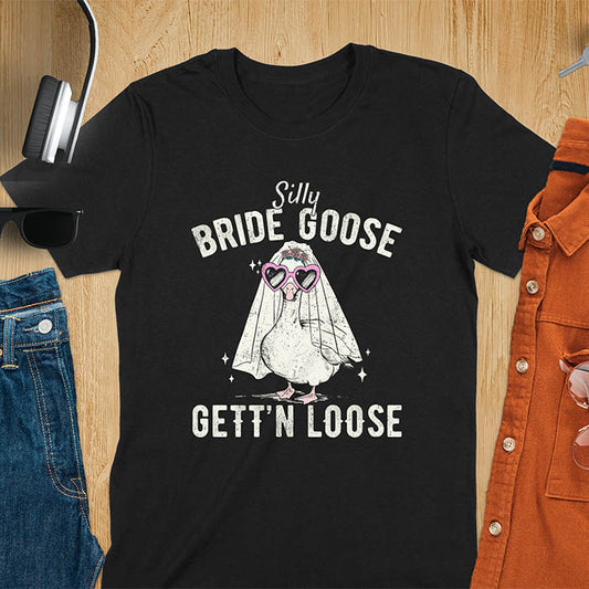 A t-shirt with a goose dressed as a bride with heart sunglasses and a veil. Text: "Silly BRIDE GOOSE" above and "GETT'N LOOSE" below. This funny bride shirt features a silly goose for getting ready bridal. A bride crewneck and engaged gift, a fiancé gift for honeymoon outfit or honeymoon gifts. A married couple shirt or new bride shirt, a fun engagement announce idea. A couple's tee-shirt and funny goose gift.