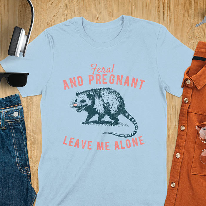 An upset-looking opossum showing its fangs. Text: "Feral AND PREGNANT LEAVE ME ALONE". This funny maternity tee for pregnancy humor is ideal for expecting mom gift & mommy to be gift. Supports feral pregnancy, pregnancy announce, expecting baby, pregnancy keepsake. A unique new mama shirt, maternity apparel & gift mom hers.