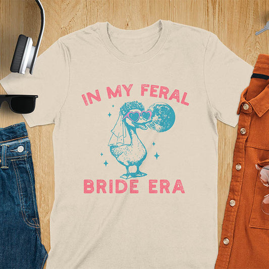 A goose standing, wearing heart-shaped sunglasses and a bride's veil. Moon in the upper right background. Retro-style text reads In My Feral Bride Era and Bride Era. Whimsical image.